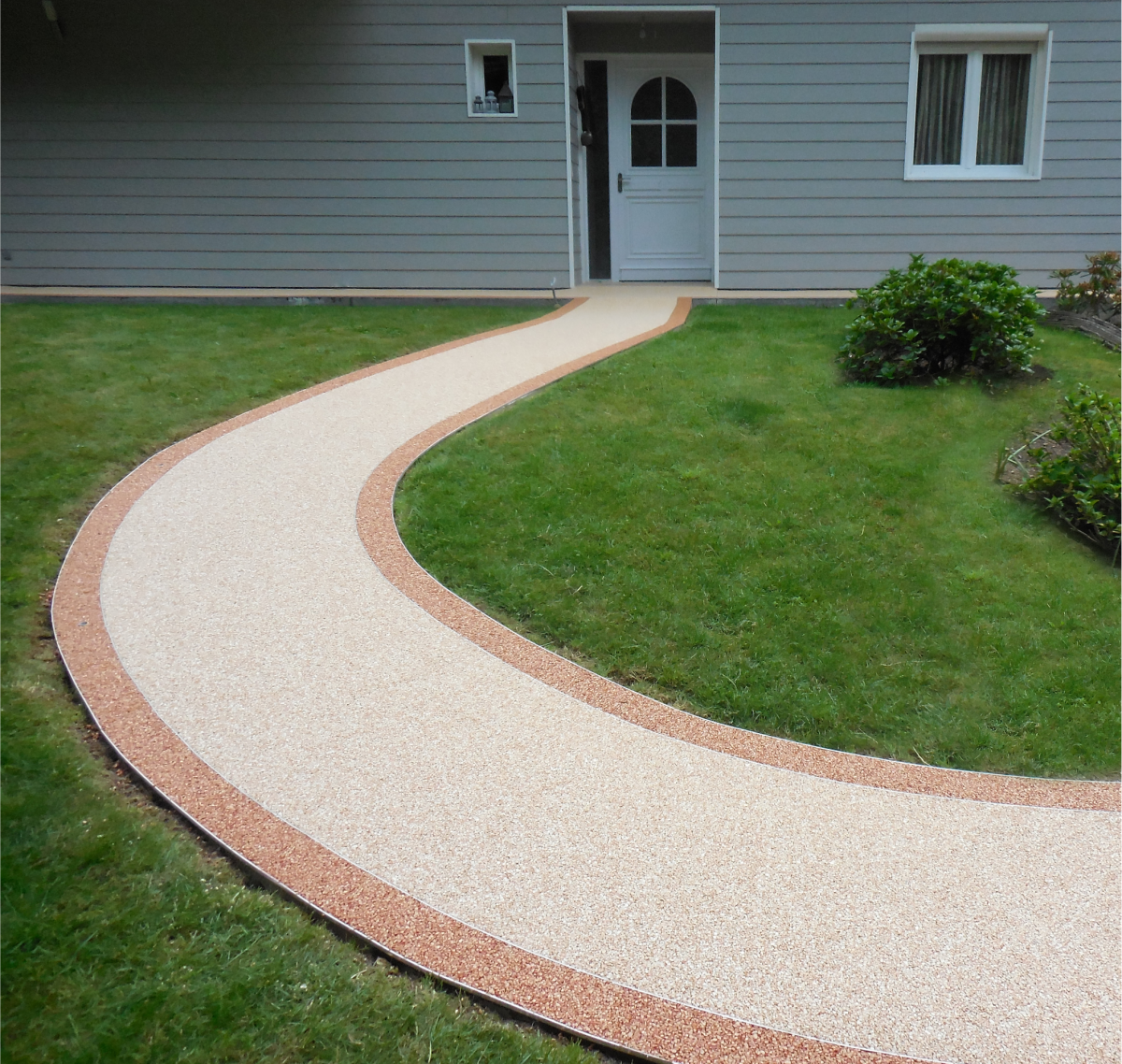 Allée courbe en moquette de pierre claire bordée d’une bande orangée sur une pelouse