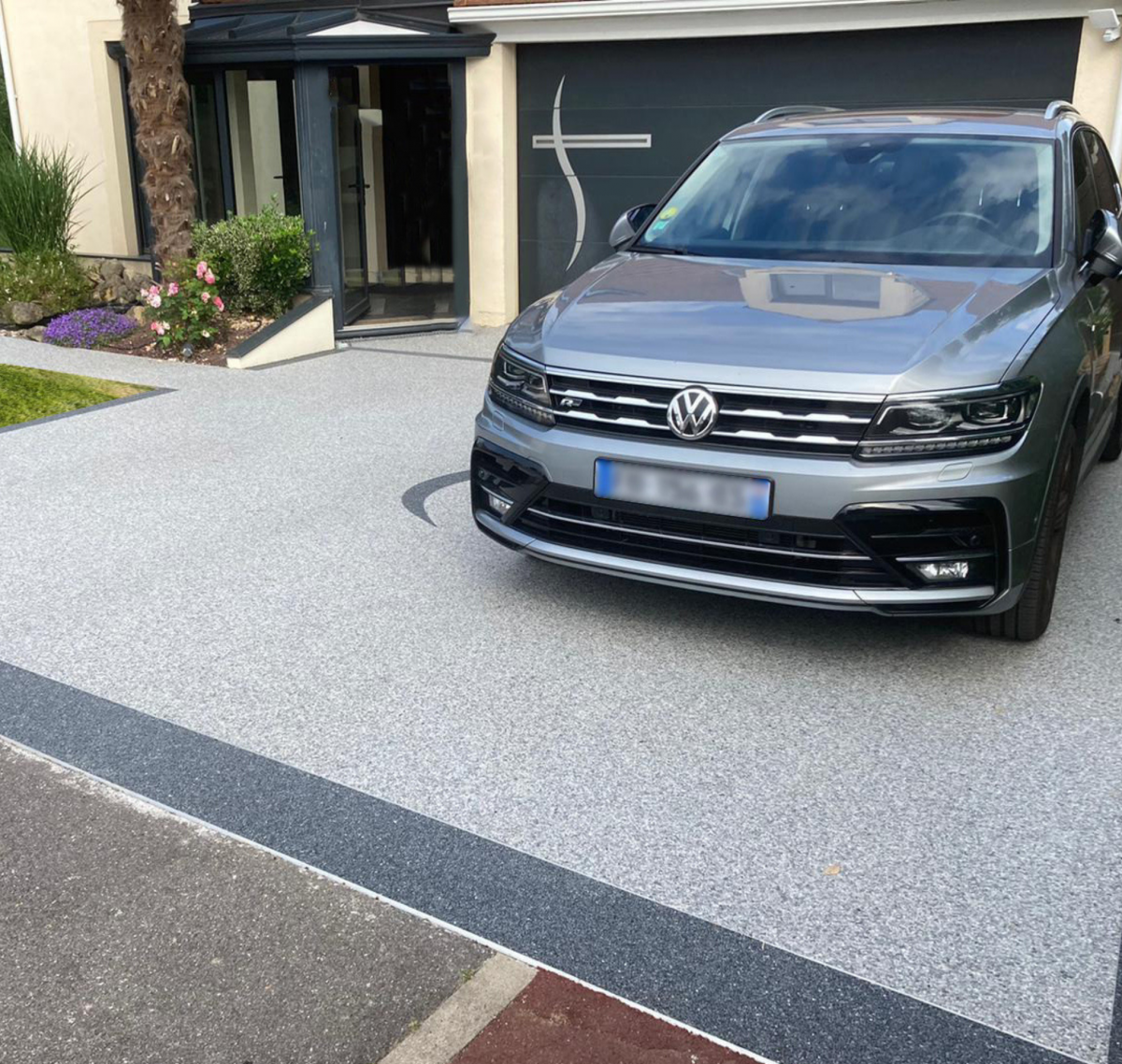 Entrée de garage carrossable en moquette de pierre grise bordée d’une bande foncée, voiture garée dessus